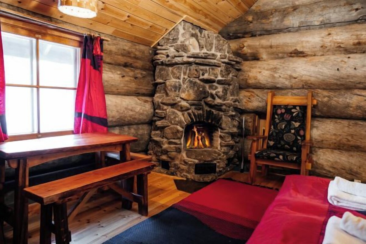 A log cabin interior with a stone fireplace, wooden furniture, and a window with red curtains.