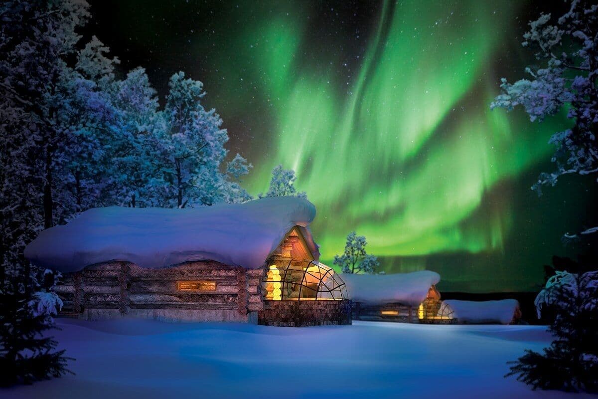 Lumihunnun peittämä hirsihökkelö, jonka katto on lumen peitossa, ja jonka ympärillä kohoavat lumipeitteiset puut. Taivaalla loistaa yöllinen revontuli.