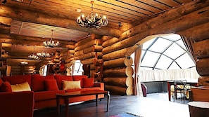 A log cabin interior with a large red sofa, wooden ceiling beams, chandeliers, and a large window.