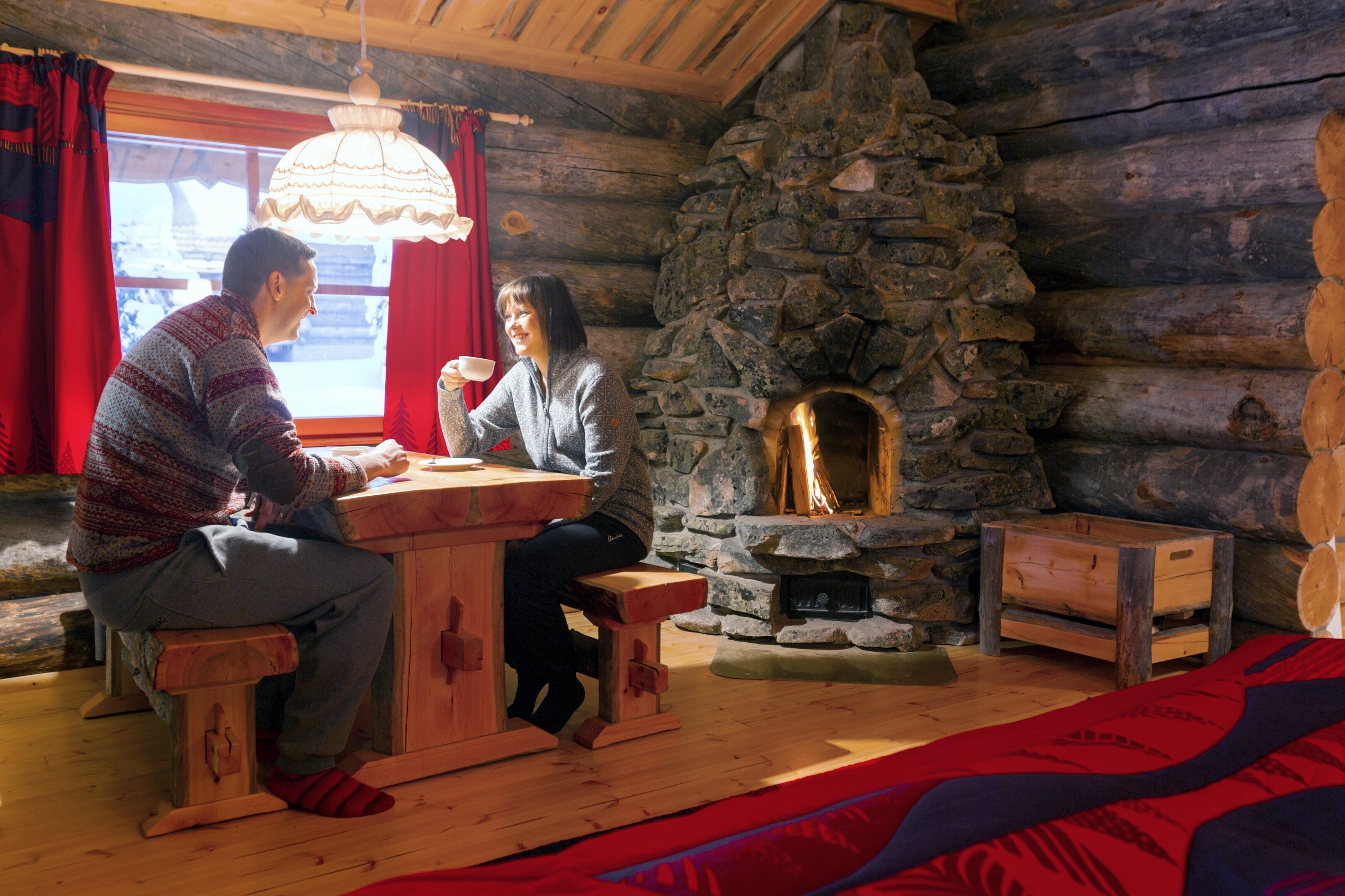 A cozy log cabin interior with a stone fireplace, wooden furniture, and a large window with red curtains.