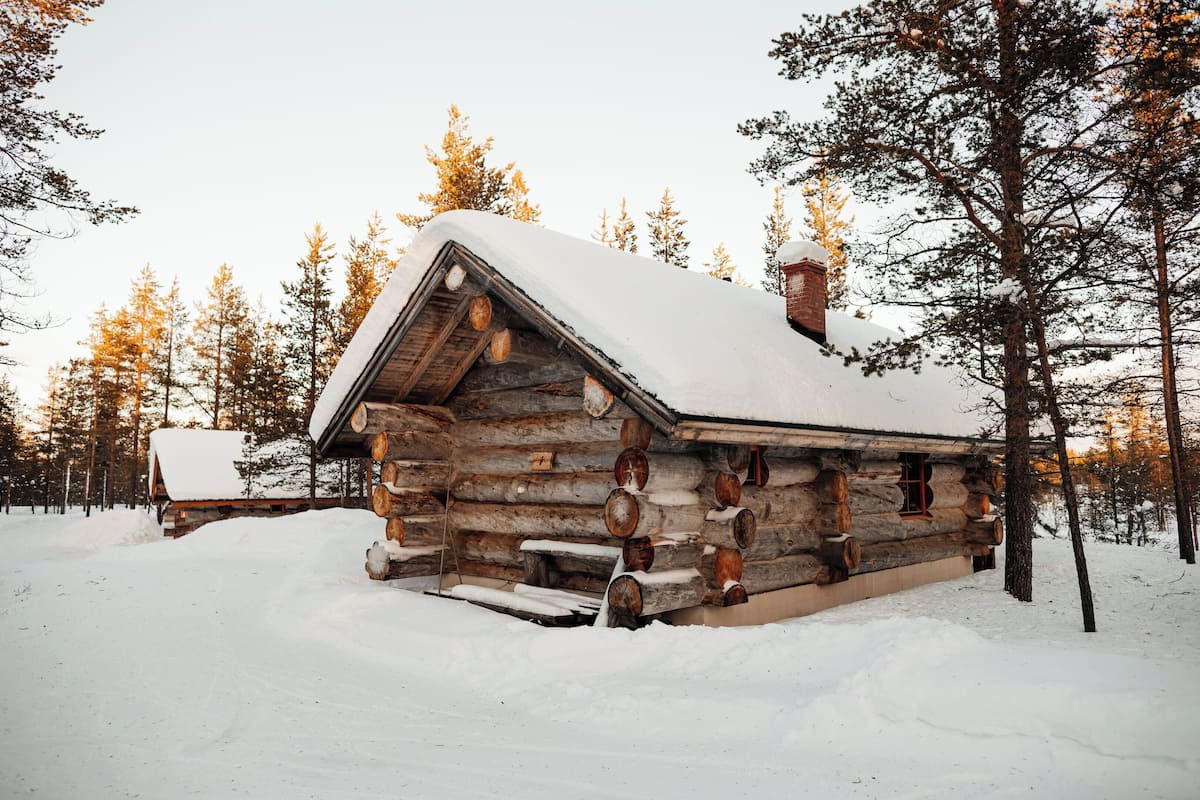 Lumihunnun peittämä hirsimökki, jonka ympärillä on puita.