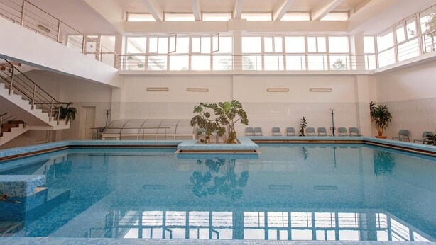 Una piscina interior con vista despejada al techo y a las ventanas, una escalera a la izquierda y una zona de estar con sillas y plantas a la derecha.