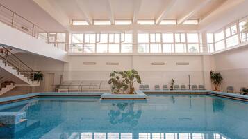 Ein Hallenbad mit freier Sicht auf Decke und Fenster, eine Treppe links sowie eine Sitzgruppe mit Stühlen und Pflanzen rechts.