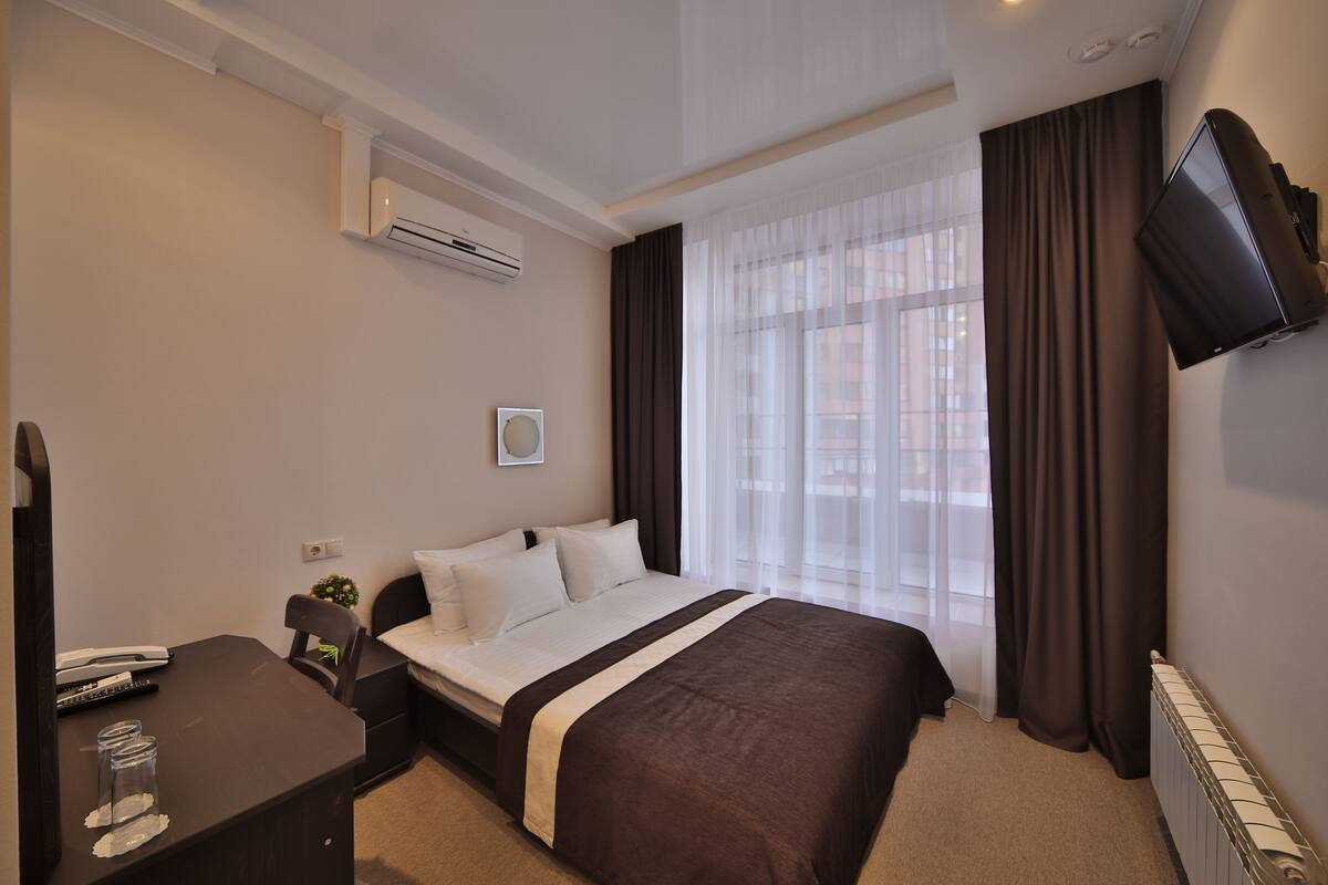 Een hotelkamer met een bed, een bureau met stoel, een airconditioning, een flatscreen televisie aan de muur en een raam met gordijnen.