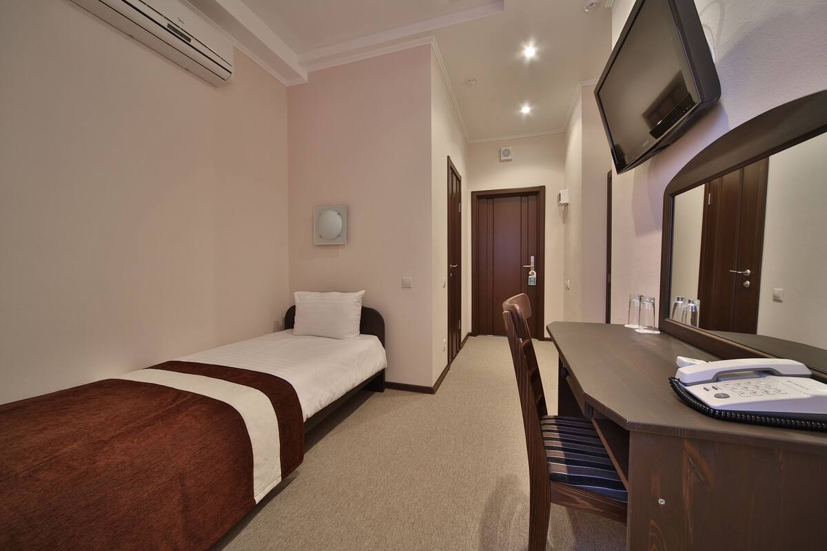 Hotelkamer met een enkel bed, een bureau met telefoon, een aan de muur bevestigde televisie en een spiegel boven het bureau.