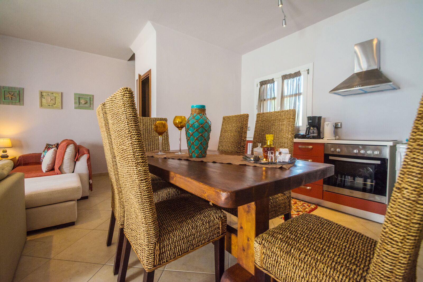 A dining area with a wooden table, wicker chairs, a vase, and a kitchen with an oven and a coffee maker.