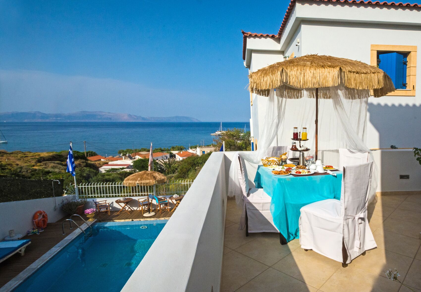 A balcony with a pool, a table set for breakfast, and a view of the sea and distant mountains.