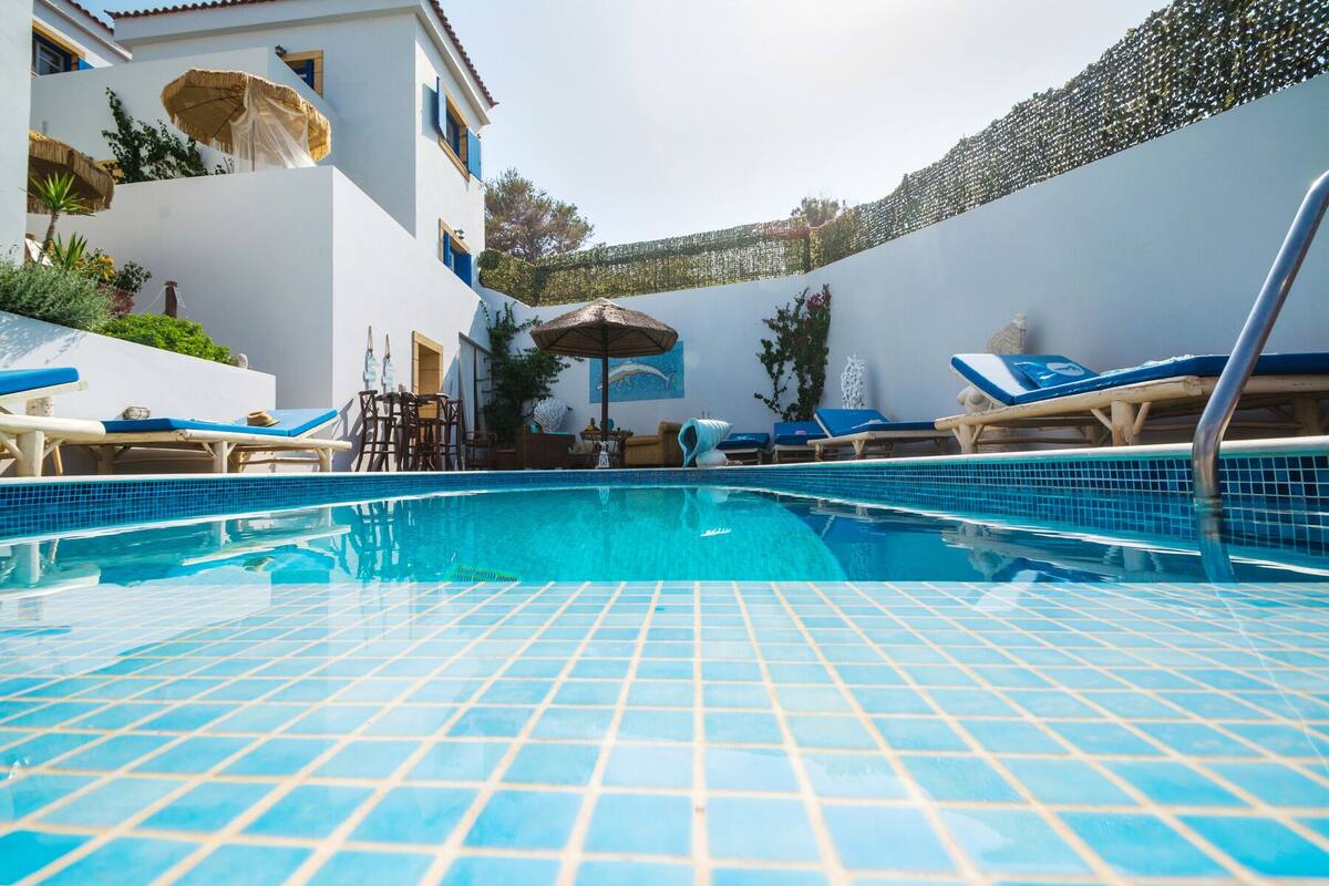 A swimming pool with tiled flooring, lounge chairs, and umbrellas.