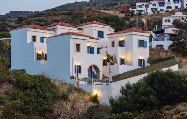 A coastal building with multiple stories, featuring a mix of white and light blue facades, red-tiled roofs, and small balconies.