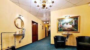 A hotel hallway with a blue carpet, a large mirror, a painting, and two black armchairs.