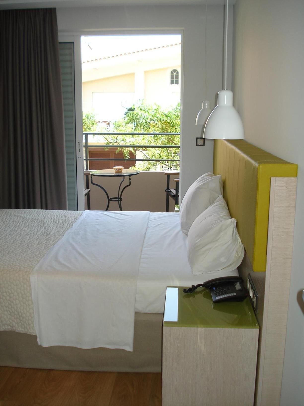 Une chambre d’hôtel avec un lit, une table de chevet avec une lampe, un téléphone et une vue sur un balcon équipé d’une table et de chaises.