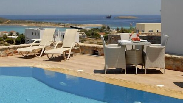 Piscina azul cristalina con sillas de descanso y una mesa, con vistas panorámicas del mar y islas lejanas.