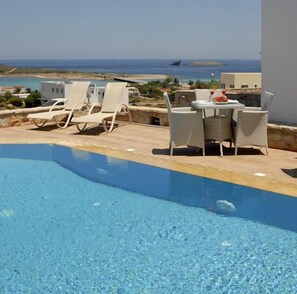 Piscina azul cristalina con sillas de descanso y una mesa, con vistas panorámicas del mar y islas lejanas.
