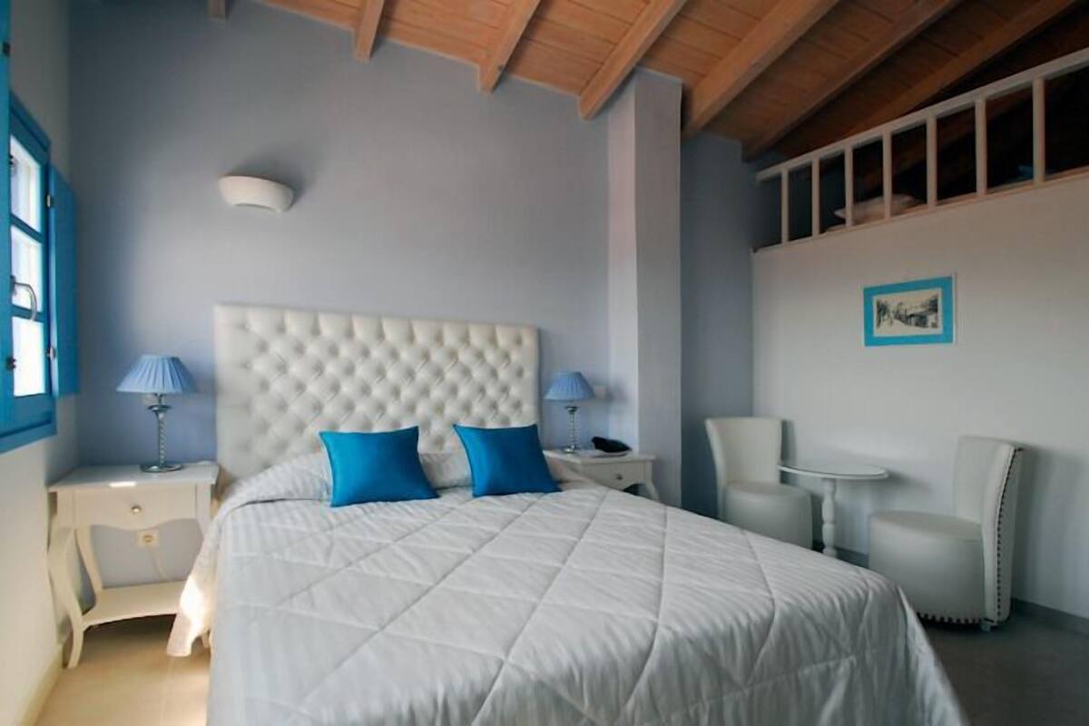 A bedroom with a large bed, two nightstands, a chair, a small table, and a window with blinds.