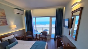 A hotel room with a bed, a desk, a chair, and a large window with a view of the beach.