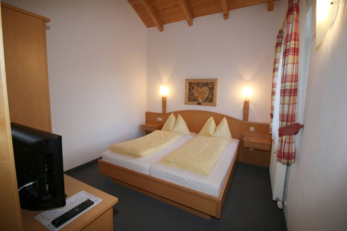 A hotel room with a wooden bed, two bedside tables, a television, and a wall-mounted light fixture.