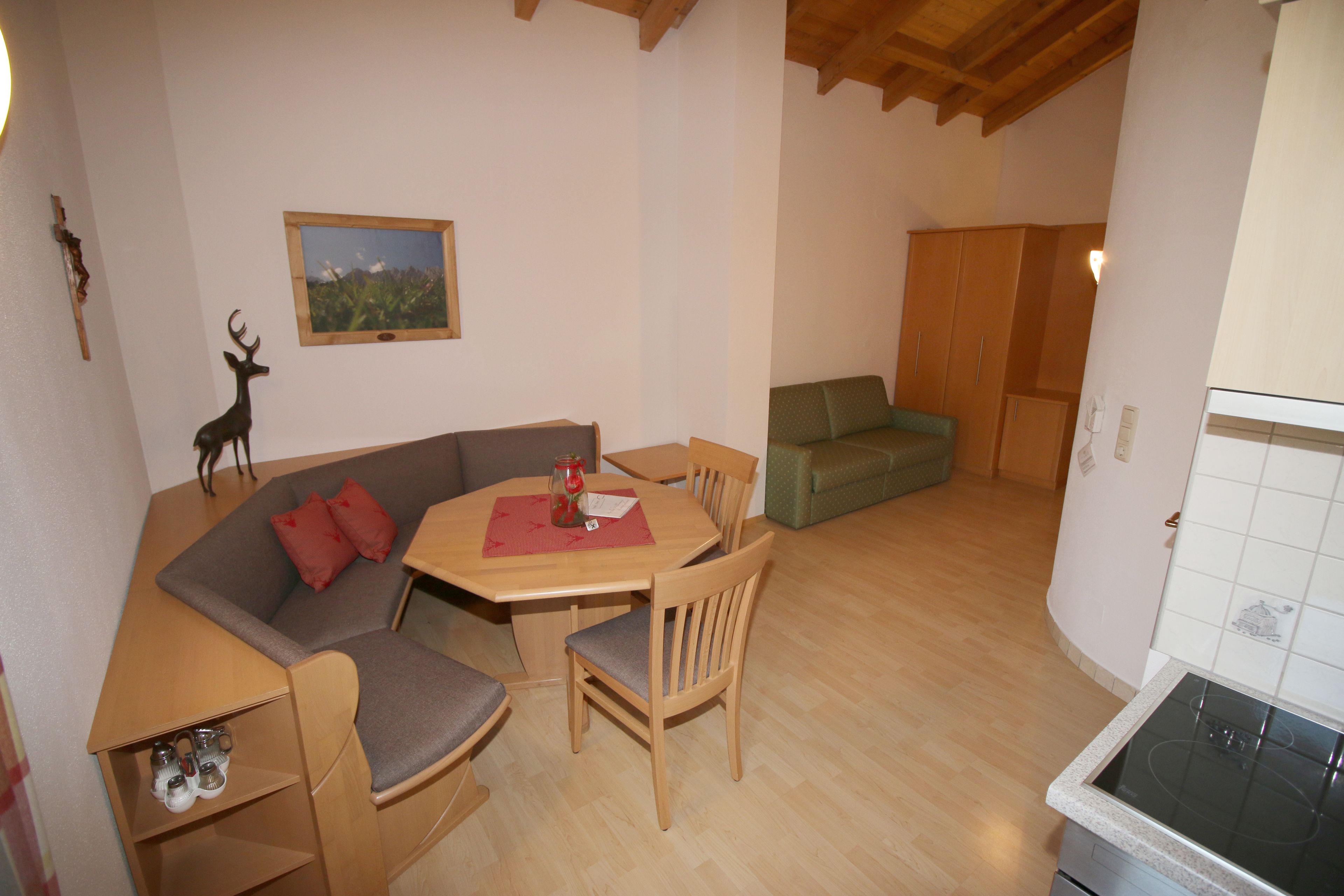 A cozy living area with a wooden dining table, a gray sofa, and a green armchair. A wooden cabinet and a kitchen area with a stove are also visible.