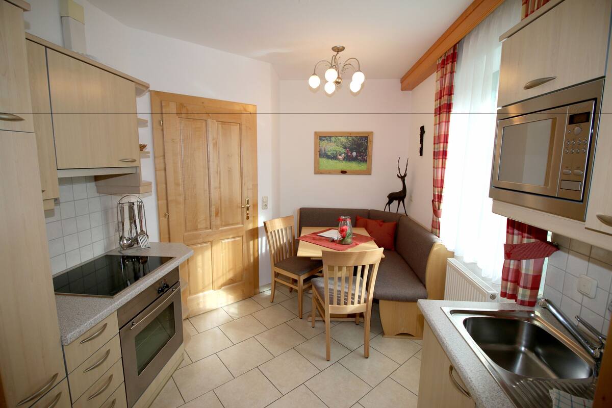 A compact kitchen with wooden cabinets, a built-in oven, a dining table with chairs, a red and white checkered curtain, and a painting on the wall.