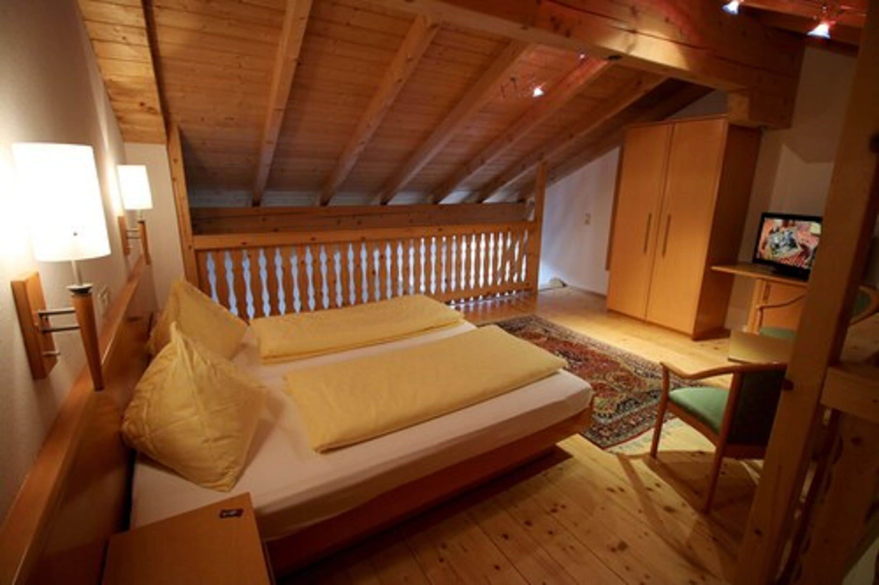 A bedroom with a wooden ceiling, a bed with yellow bedding, a small desk with a chair, and a television on a shelf.