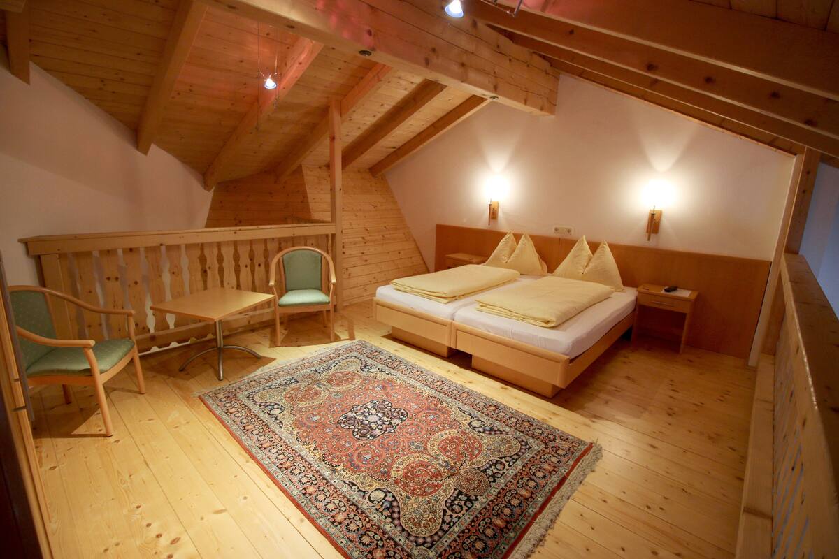 A cozy attic bedroom with a wooden floor, a bed with pillows, a nightstand, a chair, and a decorative rug.