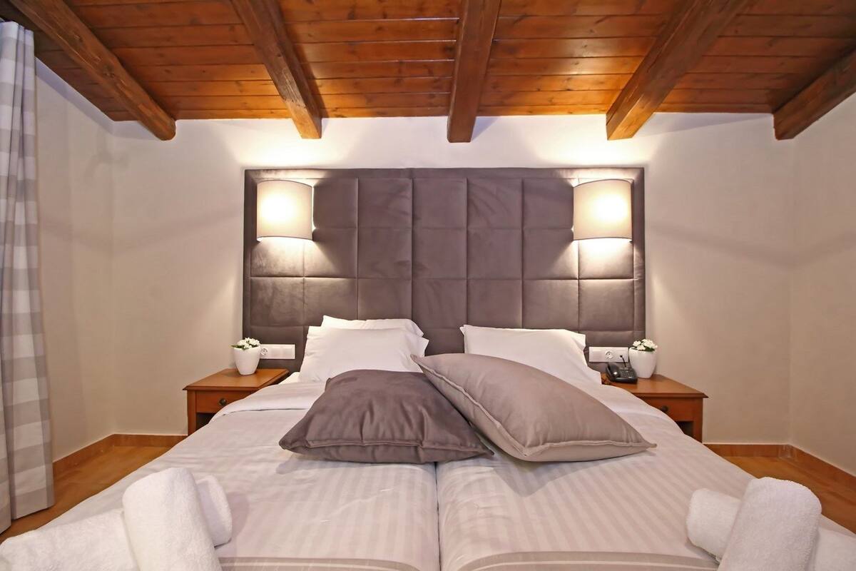 Uma cama de casal com cabeceira estofada, duas mesas de cabeceira com abajures e teto de madeira.
