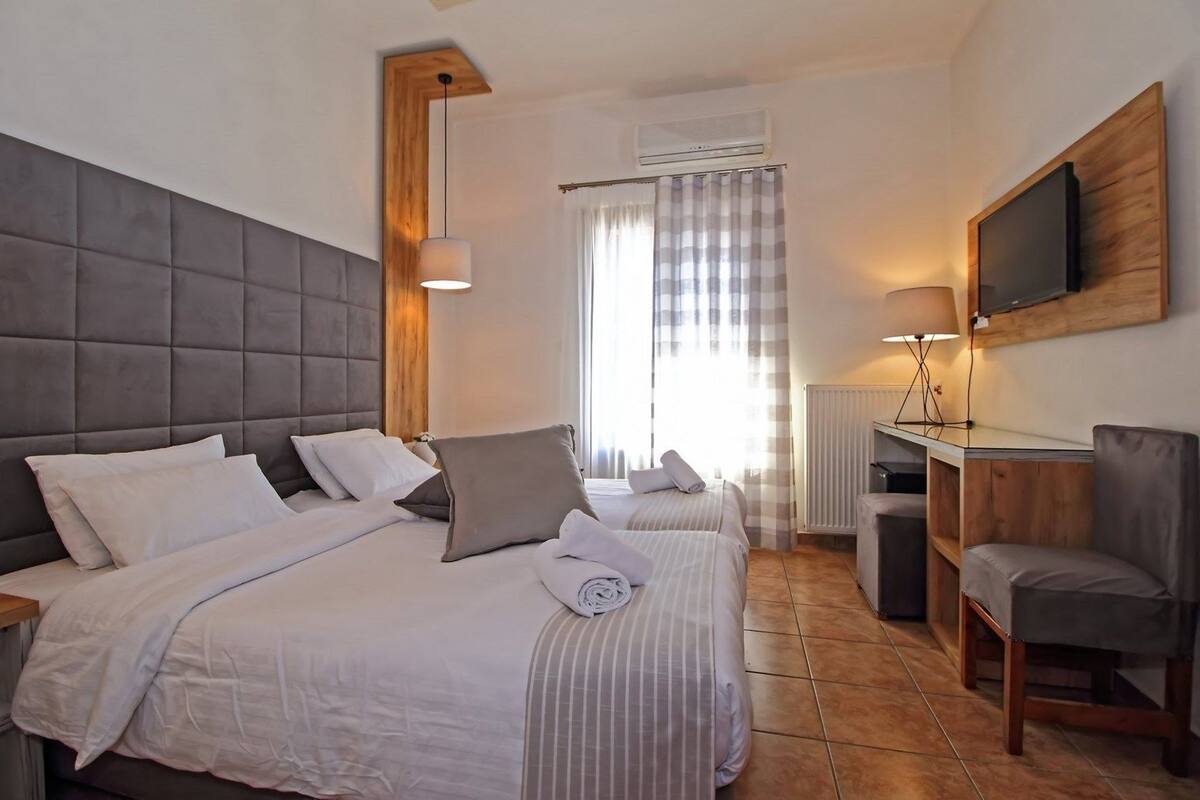 Quarto de hotel com uma cama grande, uma escrivaninha com cadeira, televisão na parede e uma janela com cortinas.