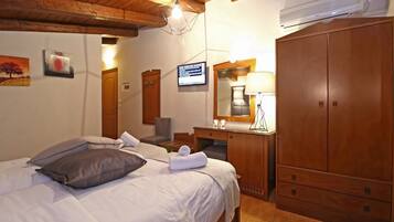 A hotel room with a wooden ceiling, a bed with pillows, a desk with a lamp, a wardrobe, and a TV on the wall.
