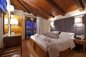 A hotel room with a wooden ceiling, a large bed with pillows and a blanket, a bedside table with a lamp, and a view of the outside through a glass door. - Arion Hotel (Delphi)