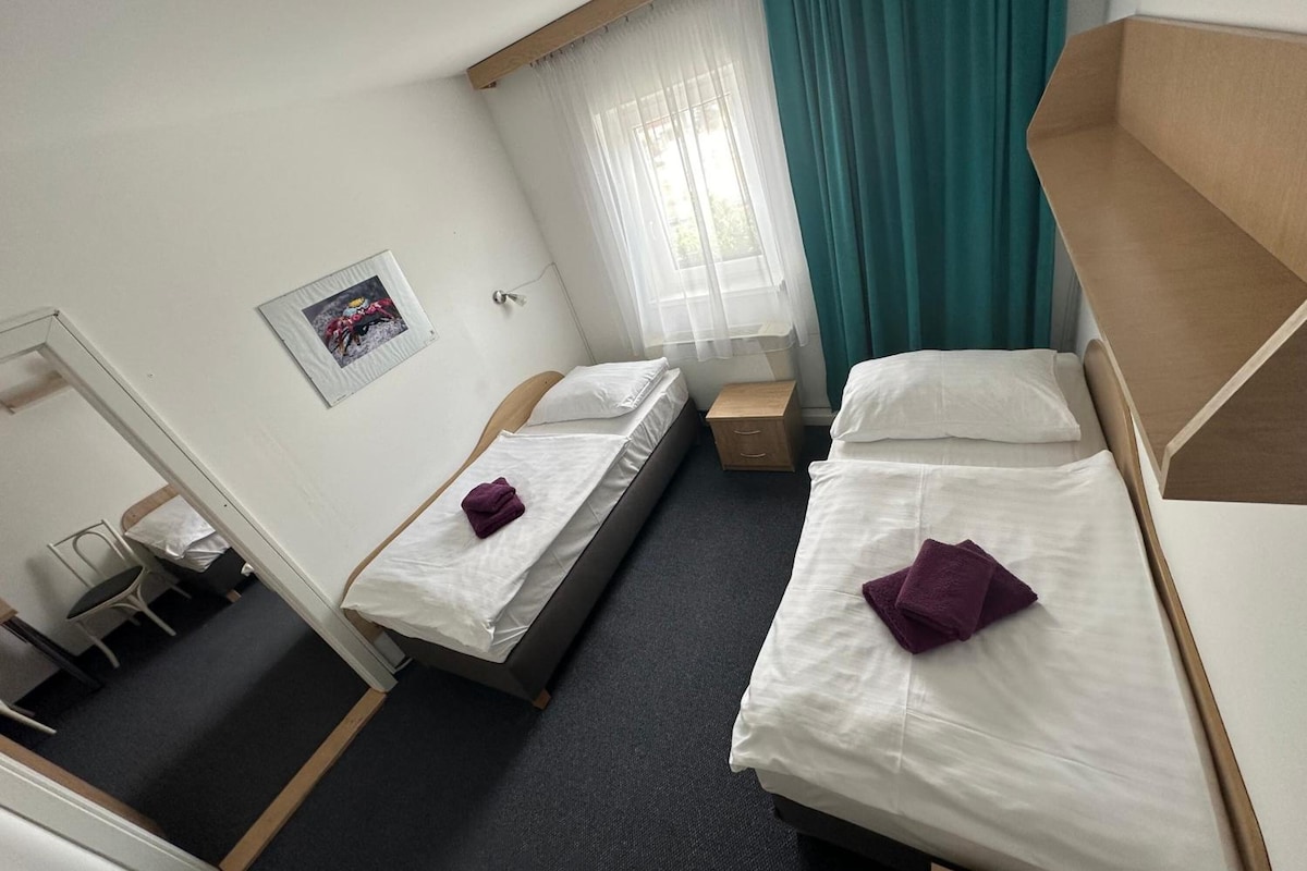 A hotel room with two single beds, a small wooden nightstand, a chair, a desk, and a window with curtains.