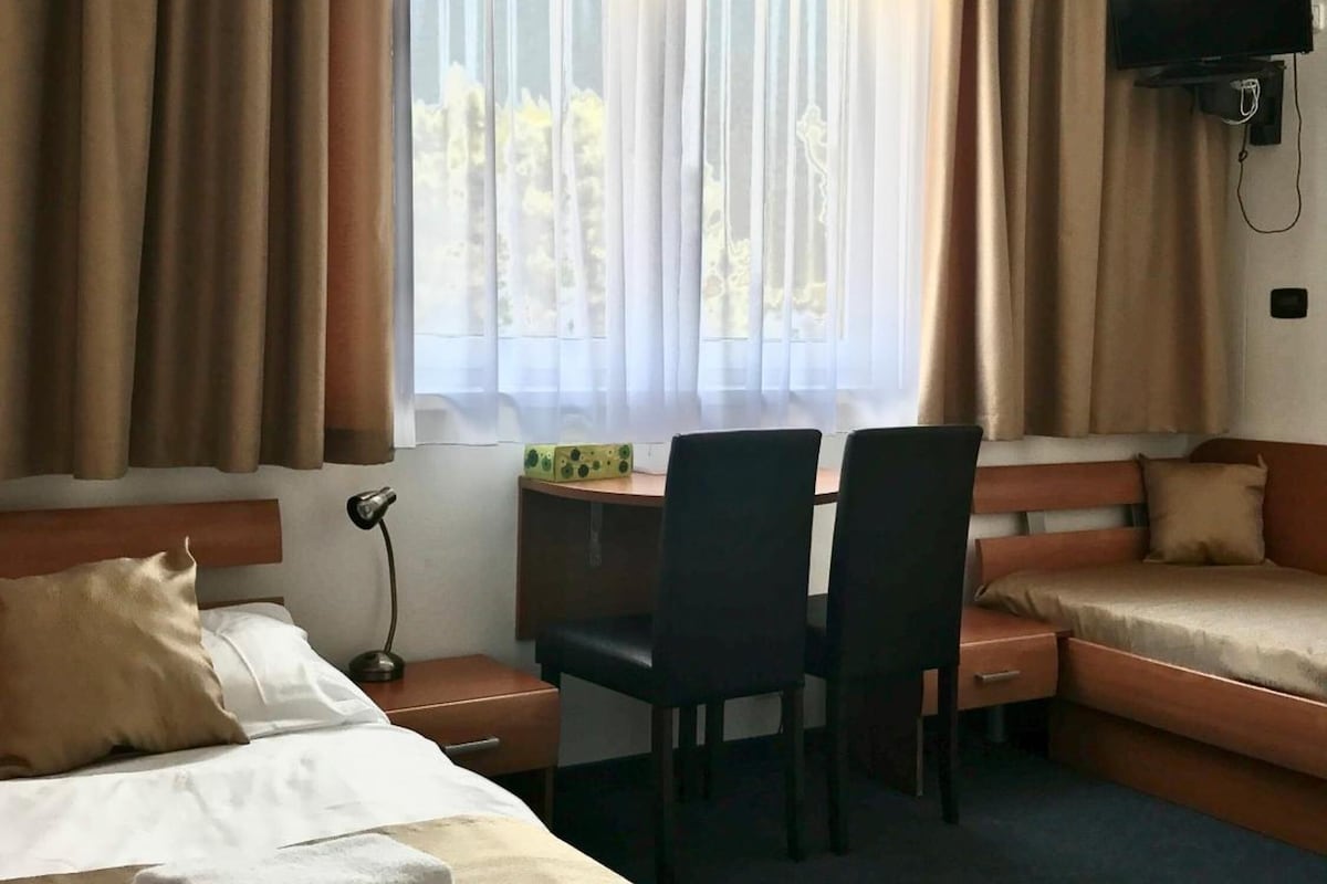 A hotel room with a bed, a desk with two chairs, a television, and a window with curtains.