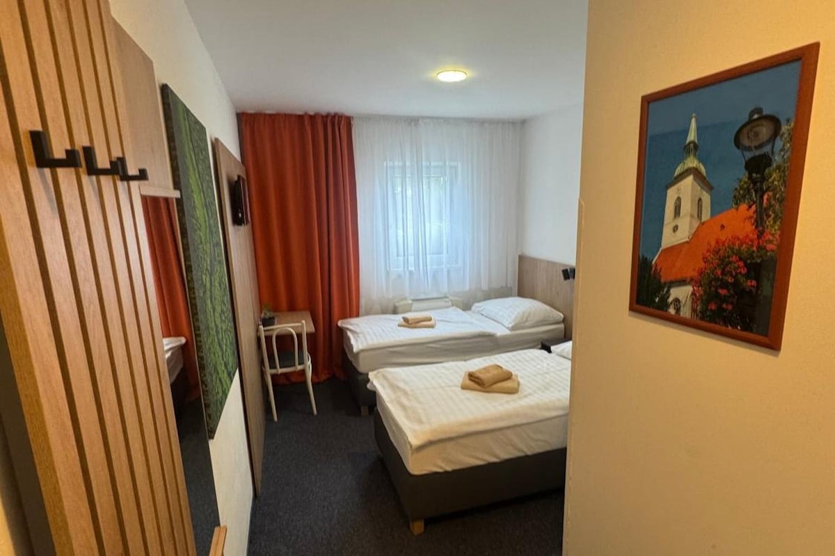 A hotel room with two beds, a small table, a chair, a painting on the wall, and a window with curtains.