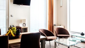 Habitación de hotel moderna con televisión de pantalla plana, una mesa de comedor con sillas, frutero y un gran ventanal con cortinas.