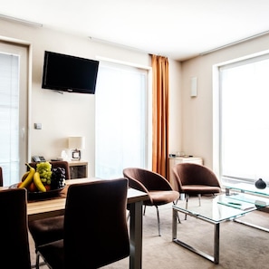 A modern hotel room with a flat-screen TV, a dining table with chairs, a fruit bowl, and a large window with curtains. - Deluxe (Nitra)