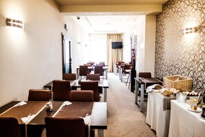 A hotel dining area with multiple tables and chairs, a television screen, and a patterned wall.