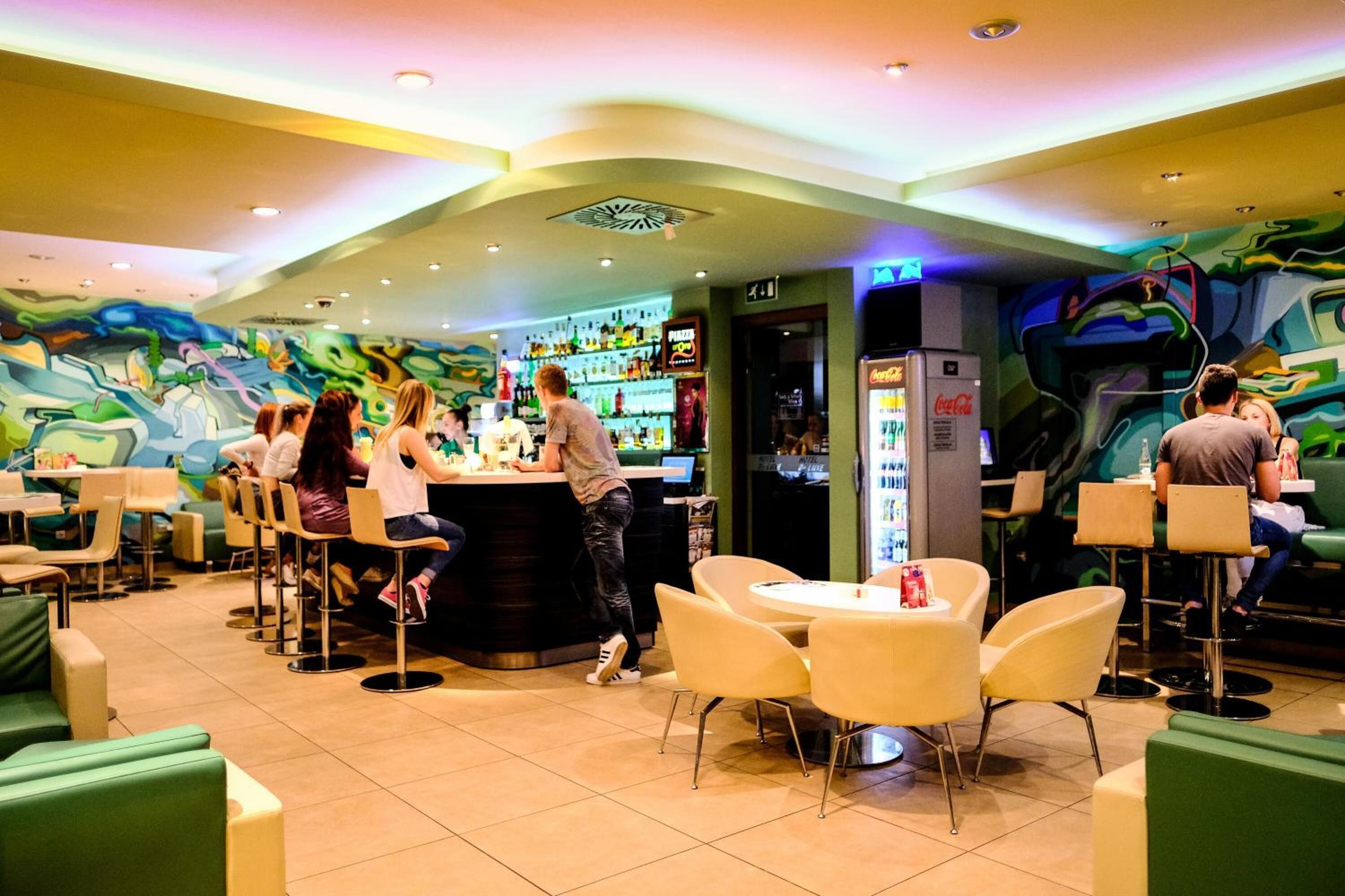 Un restaurante con zona de bar, disposición de mesas y un mural colorido en la pared.