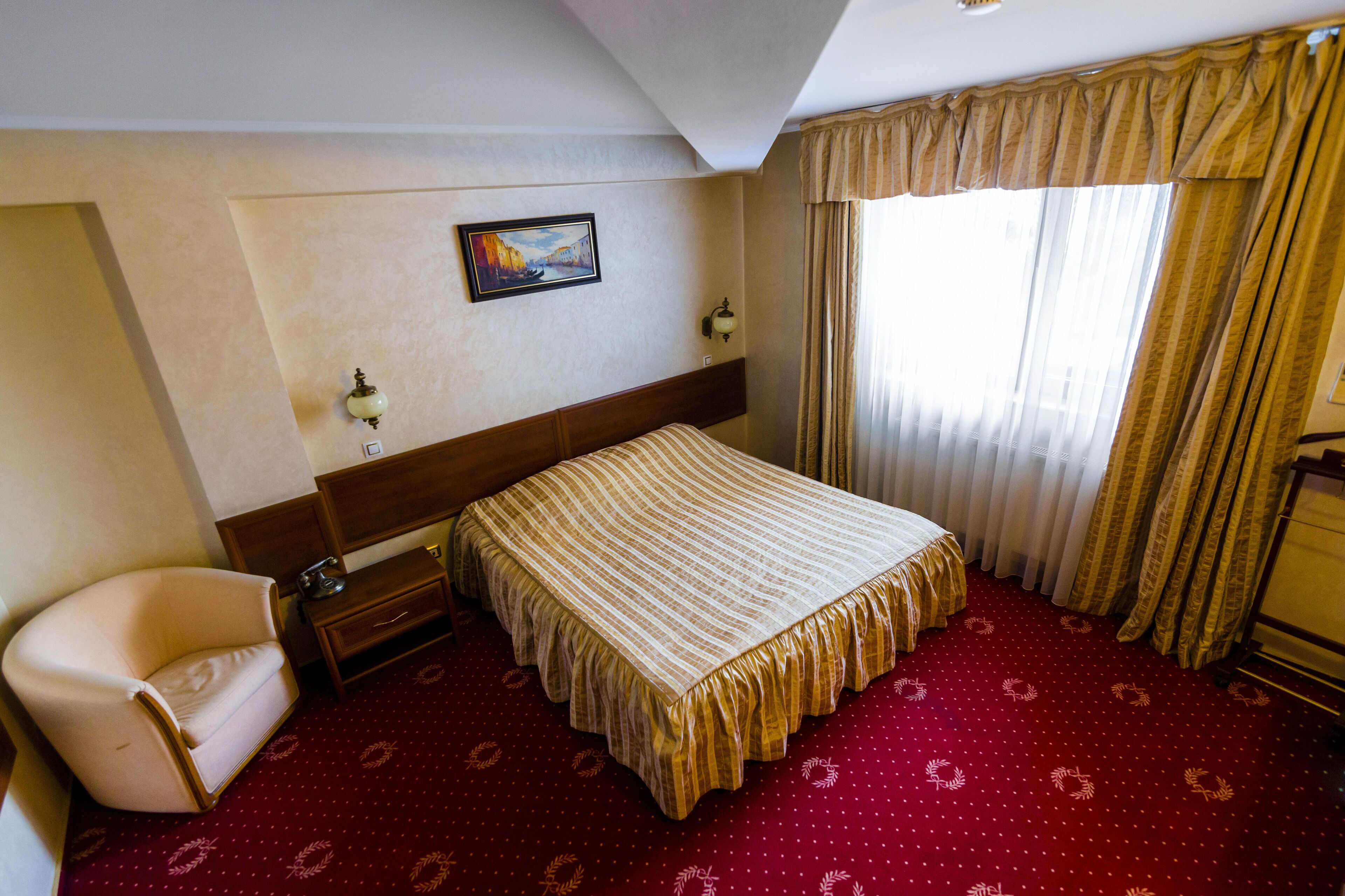 A hotel room with a bed, a chair, a nightstand, a painting on the wall, and a window with curtains.