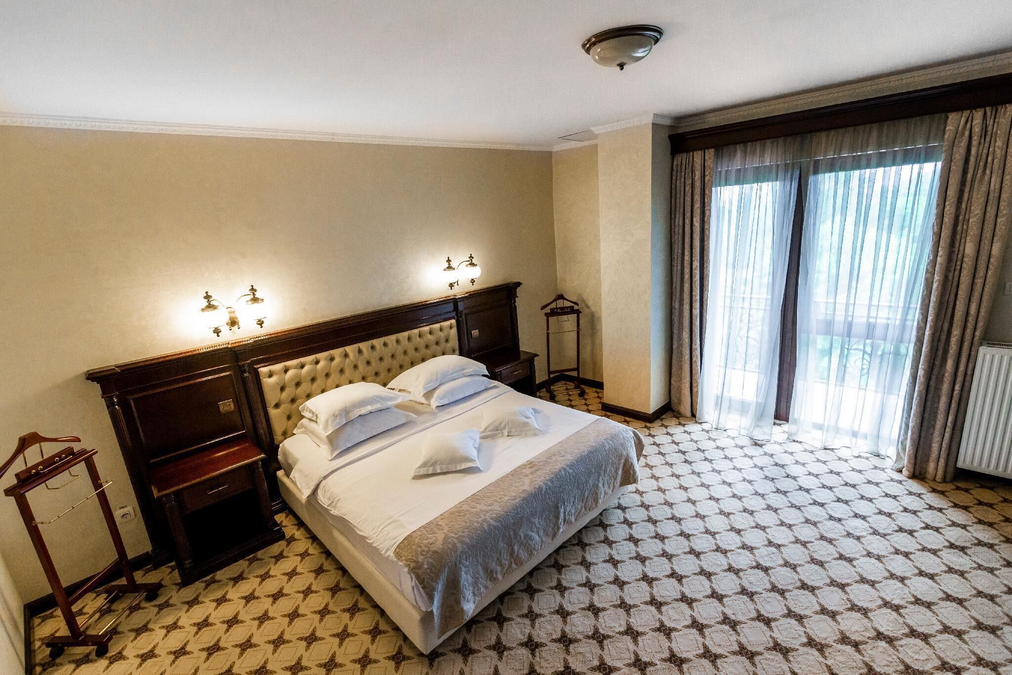 A hotel room with a large bed, a nightstand, a lamp, a wardrobe, and a window with curtains.