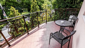 A balcony with a table and chairs, a metal railing, and a view of trees and a road.