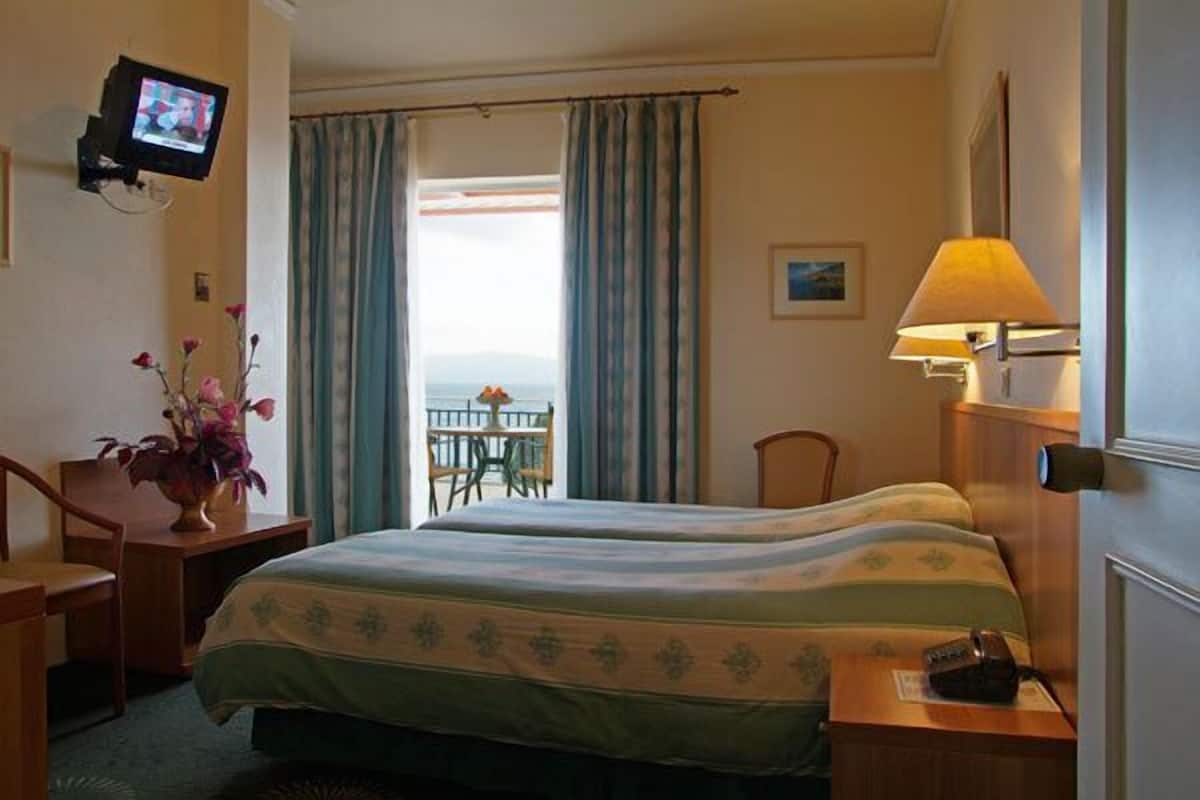 Habitación de hotel con dos camas, un televisor montado en la pared, un escritorio con una lámpara y vista al mar por la ventana.