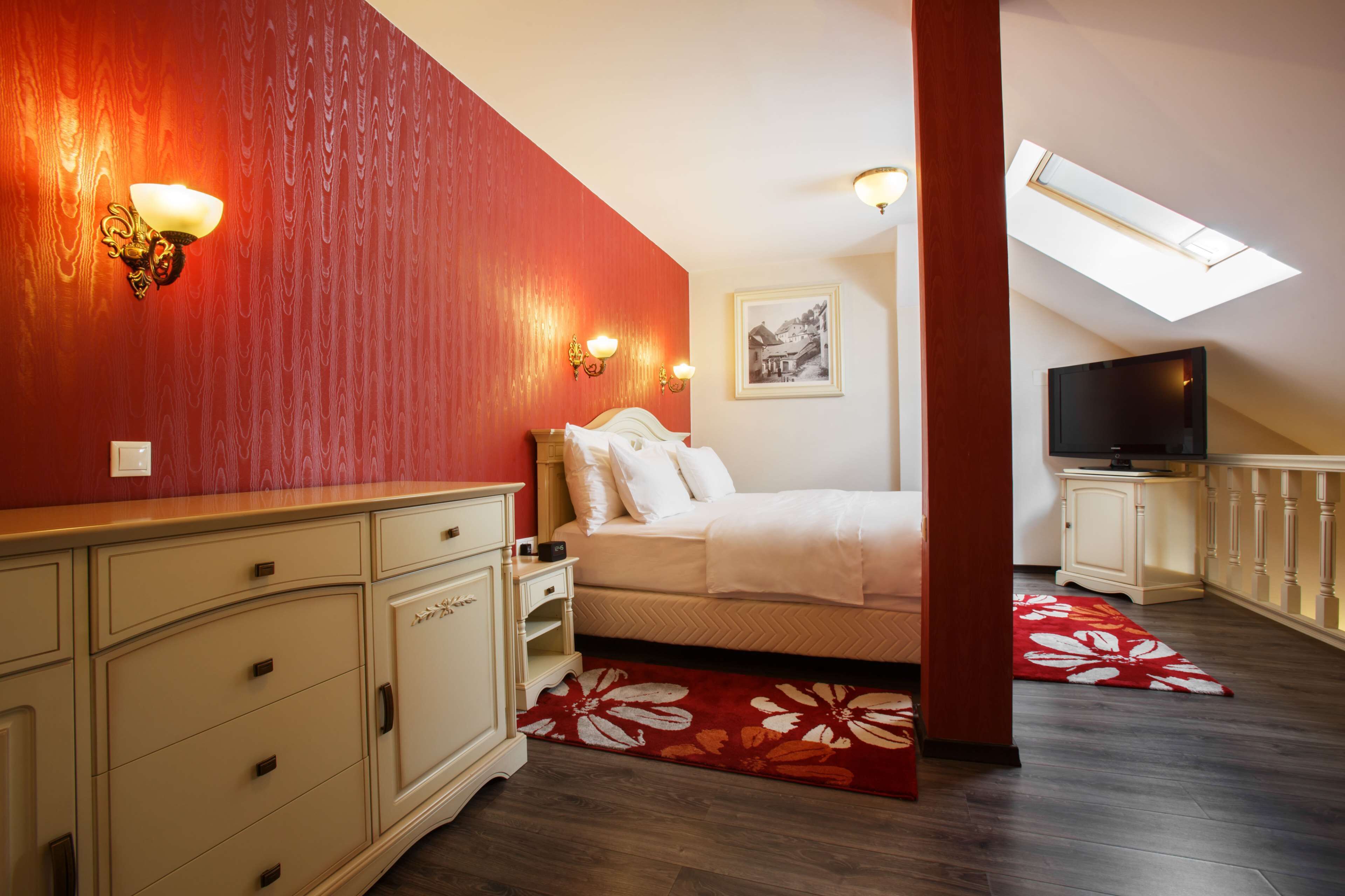 A bedroom with a bed, a TV, a red wall, and a skylight.