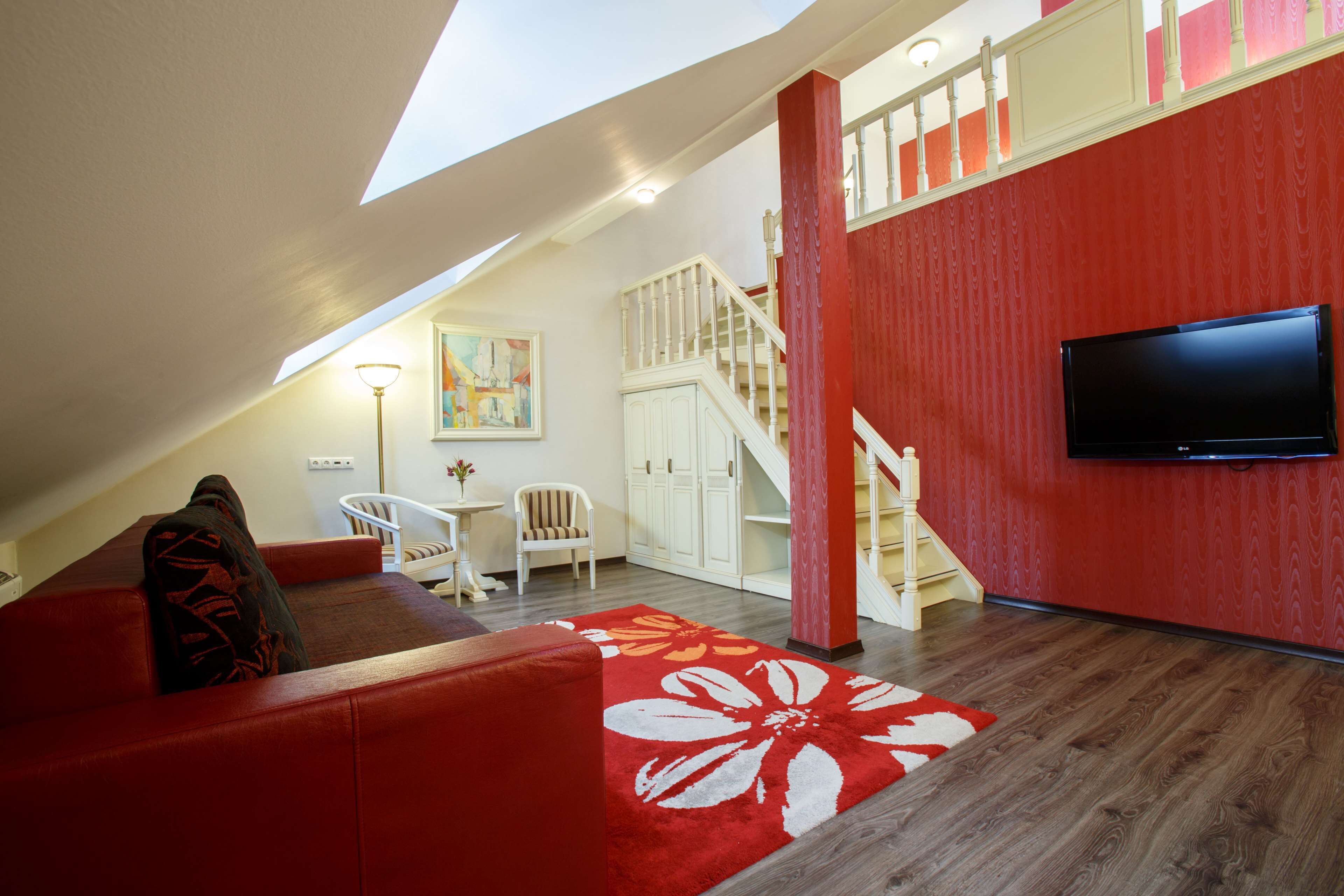 A modern living room with a red sofa, a wooden floor, a staircase, and a flat-screen TV mounted on the wall.