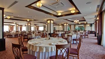A spacious banquet hall with round tables set for dining, featuring wooden chairs and white tablecloths.