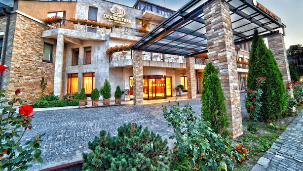 Un hotel con fachada de piedra, entrada de vidrio y un balcón con flores rojas.