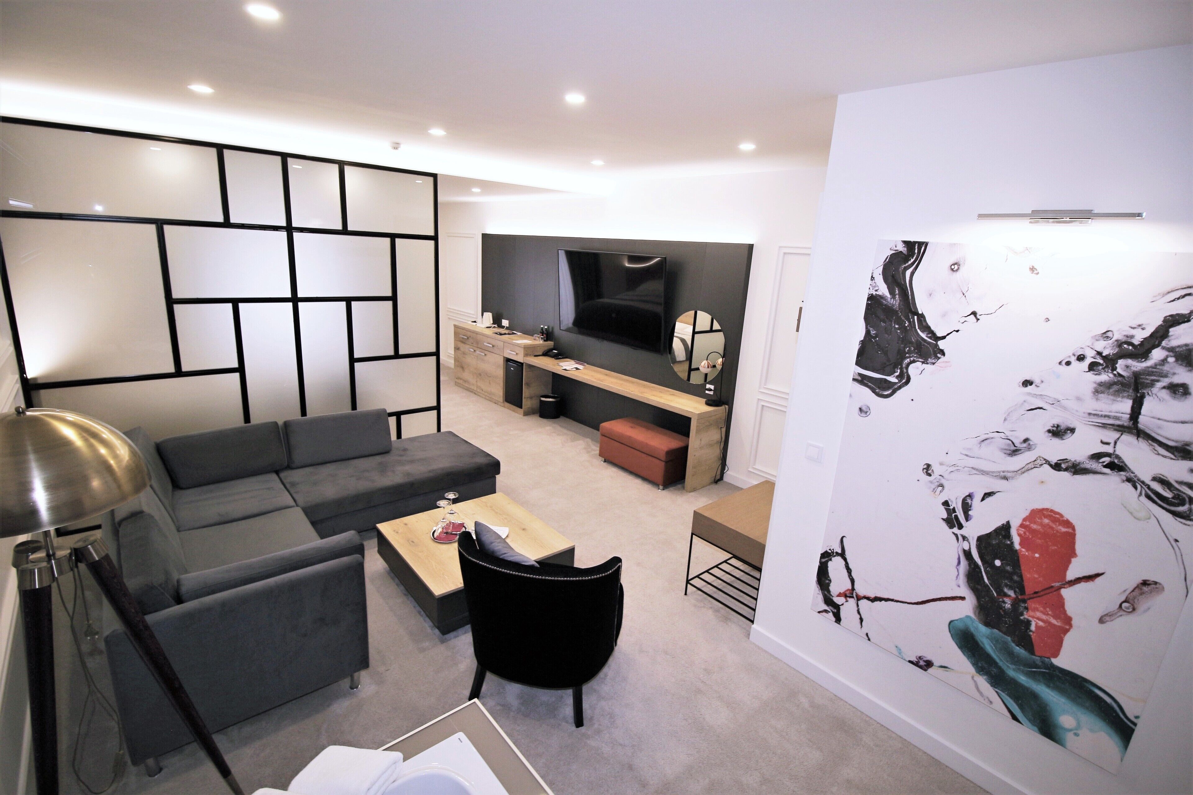 Un salon moderne avec un canapé d’angle, une table basse et un fauteuil noir. Un téléviseur est fixé au mur, ainsi qu’un meuble en bois et un grand tableau abstrait.