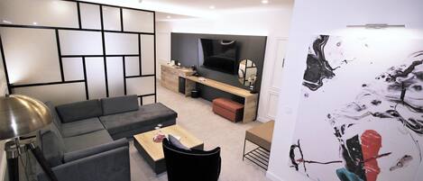 A modern living room with a sectional sofa, a coffee table, and a black chair. There is a TV mounted on the wall, a wooden cabinet, and a large abstract painting on the wall.