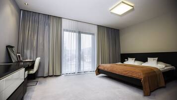 Quarto de hotel moderno com cama, escrivaninha, cadeira e grandes janelas com cortinas.