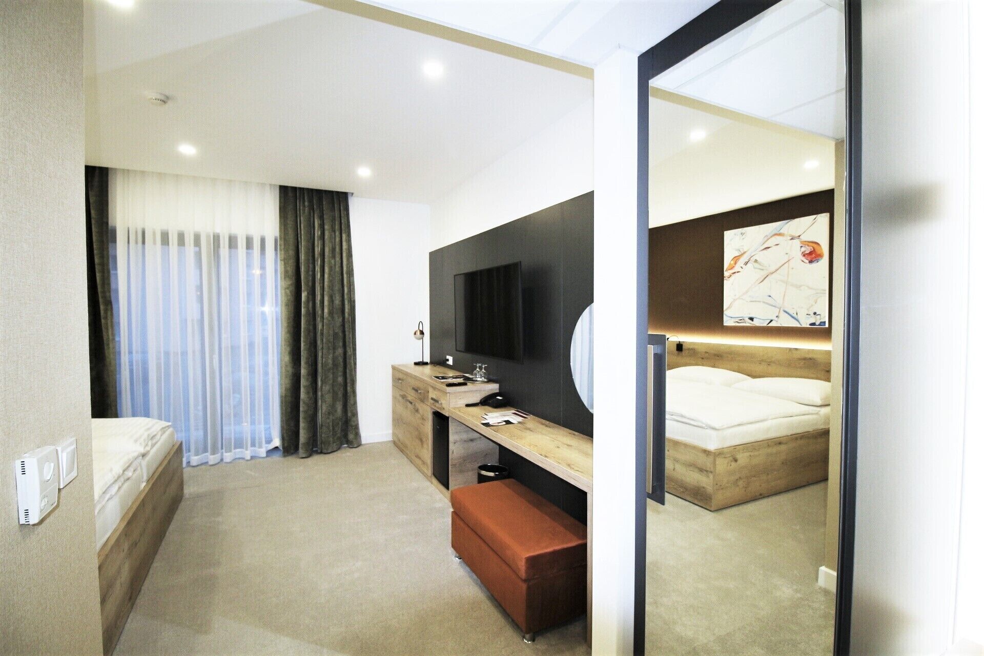 A modern hotel room with a bed, a desk with a flat-screen TV, a wooden nightstand, a small bench, and a large mirror.