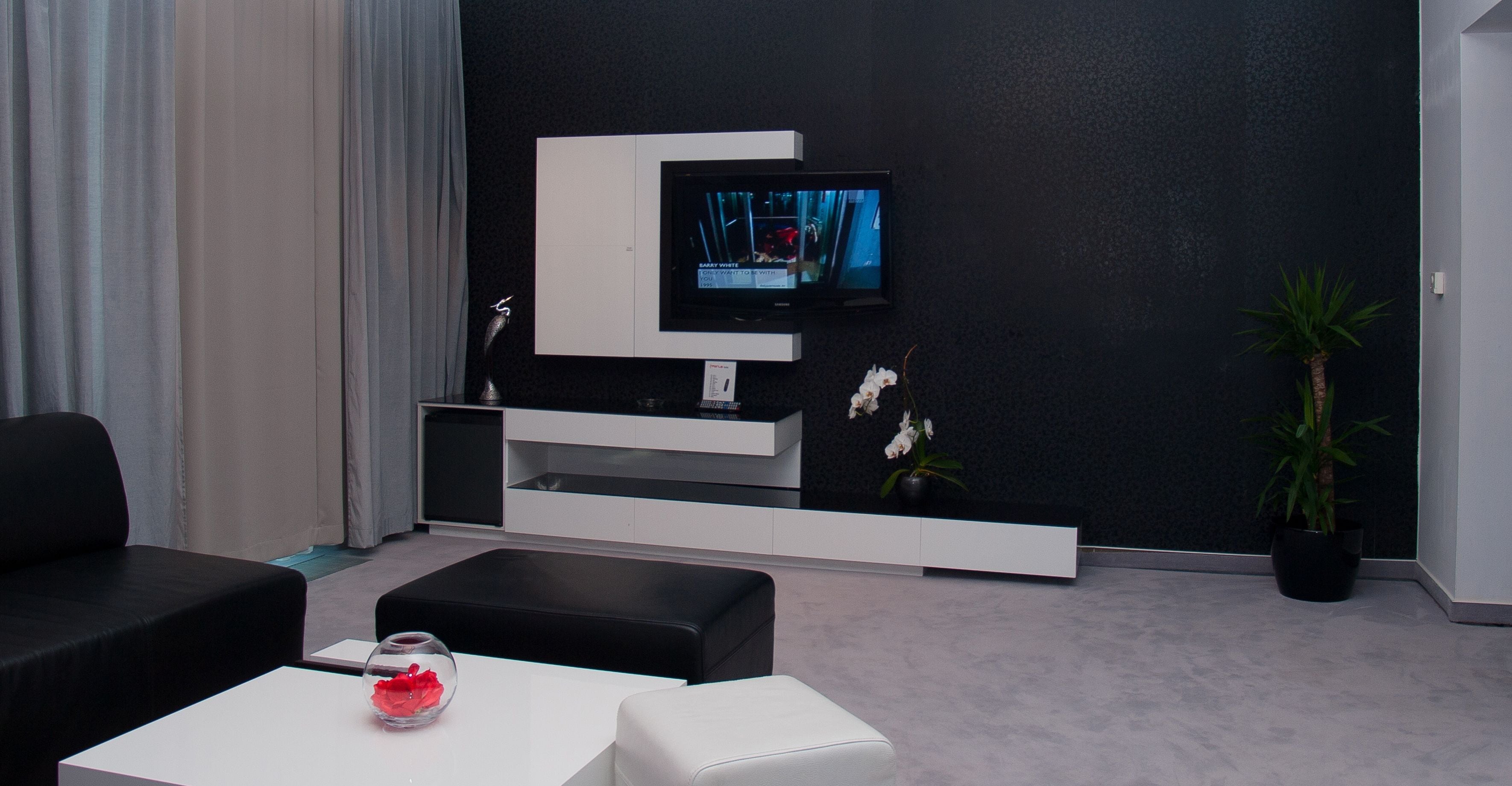 Una sala de estar moderna con un televisor de pantalla plana montado en una pared oscura, un sofá blanco, un reposapiés negro y una mesita con un jarrón de cristal.