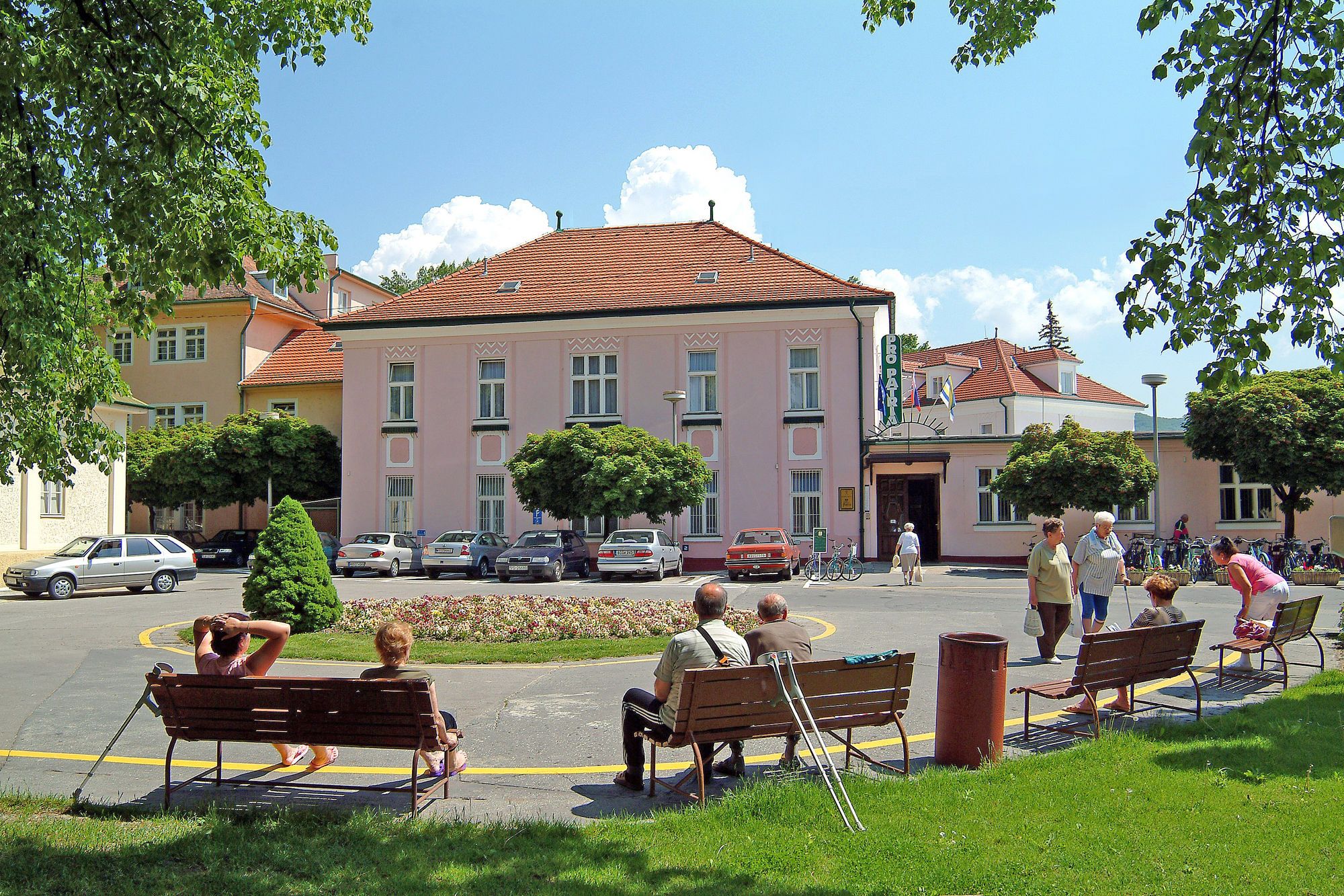 A public square with benches, a building with a red roof, and parked cars.