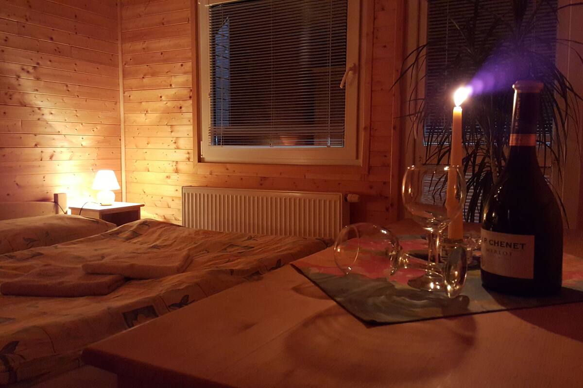 A cozy room with wooden walls, a bed with a blanket, a bedside table with a lamp, a window with blinds, and a table with a bottle of wine, glasses, and a candle.